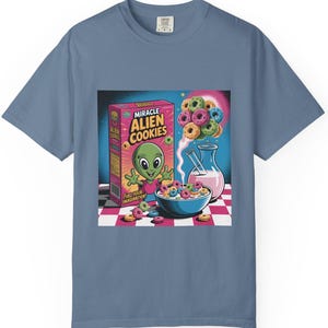 May include: Blue t-shirt featuring a colorful graphic of a box of "Miracle Alien Cookies" cereal with an alien mascot, a bowl of cereal, and a pink drink. The design includes a checkerboard pattern.
