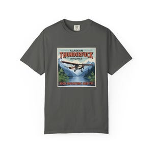 May include: Dark gray t-shirt featuring a vintage-style graphic. The graphic includes the text "ALASKAN THUNDERFUCK AIRLINES" above an image of a seaplane flying over a lake and mountains. The text "HIGH ADVENTURE AWAITS!" is below the image.