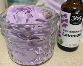 May include: A small glass jar filled with lavender-coloured cream, next to a brown glass bottle of lavender essential oil. The bottle has a white label with purple floral graphics and the text "365 Whole Foods Market".