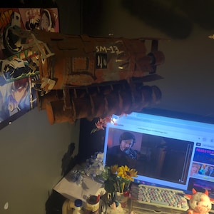 May include: A brown, cylindrical, industrial-style prop model labeled SM-13 with a textured, weathered surface and multiple protruding circular components, mounted on a wall above a computer monitor displaying a YouTube video interface.