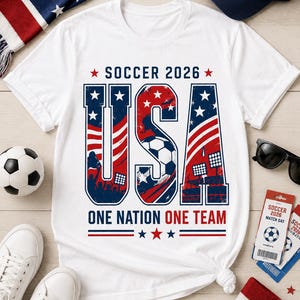 May include: White t-shirt with a patriotic design for the 2026 Soccer World Cup. The word "USA" is prominently displayed in red, white, and blue, with a soccer ball graphic. The text "SOCCER 2026" and "ONE NATION ONE TEAM" are also featured.