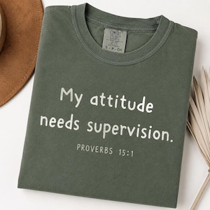 May include: An olive green crew neck t-shirt with white text that reads "My attitude needs supervision." and "PROVERBS 15:1" printed below. The shirt features a Comfort Colors brand label at the inner neckline indicating a size small.