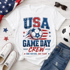 May include: White t-shirt with a graphic design featuring "USA" in red and blue, a soccer ball, and the text "GAME DAY CREW." The design includes stars and the phrase "ONE NATION. ONE TEAM."