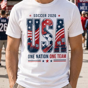May include: White t-shirt with a patriotic design for the 2026 Soccer World Cup. The shirt features the word "USA" in red, white, and blue, with a soccer ball graphic and the text "ONE NATION ONE TEAM".