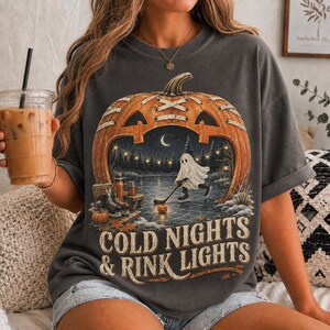 May include: A charcoal gray t-shirt featuring a large graphic of a carved pumpkin shaped like a hockey rink. Inside the pumpkin, a ghost plays hockey on ice under string lights. The text below reads COLD NIGHTS & RINK LIGHTS in a vintage font.