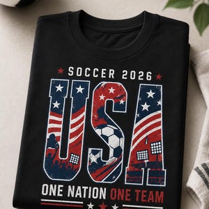 May include: Black t-shirt with a patriotic design for the 2026 Soccer World Cup. The word "USA" is prominently displayed in red, white, and blue, with a soccer ball graphic. The text "SOCCER 2026" and "ONE NATION ONE TEAM" are also featured.