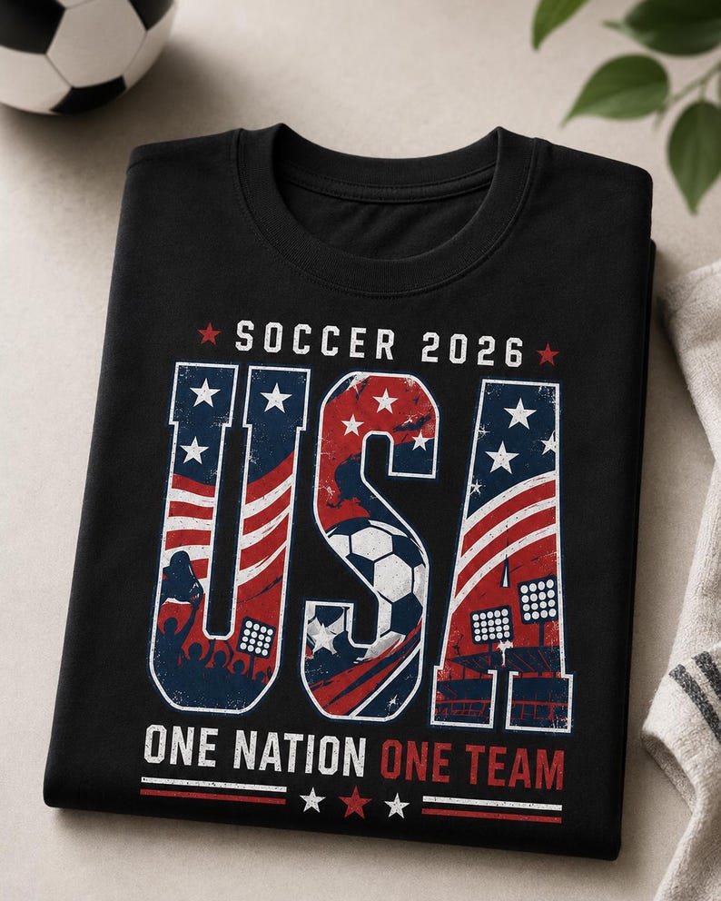 May include: Black t-shirt with a patriotic design for the 2026 Soccer World Cup. The word "USA" is prominently displayed in red, white, and blue, with a soccer ball graphic. The text "SOCCER 2026" and "ONE NATION ONE TEAM" are also featured.