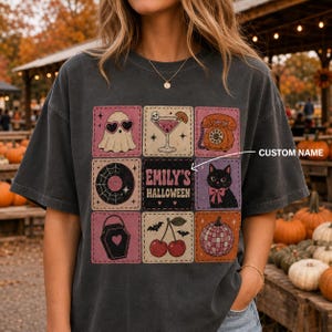 May include: A charcoal grey t-shirt featuring a grid of nine square patches with Halloween-themed illustrations including a ghost, a martini, a rotary phone, a vinyl record, a black cat, a coffin, cherries, and a pumpkin, with text reading EMILY'S HALLOWEEN.