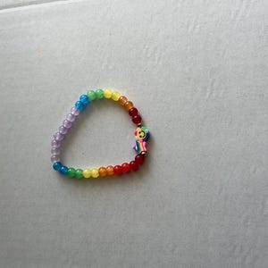 May include: A rainbow-colored beaded bracelet with a charm. The bracelet features a gradient of colors, including purple, blue, green, yellow, orange, and red beads. A small, colorful charm with a smiling face is attached to the bracelet.