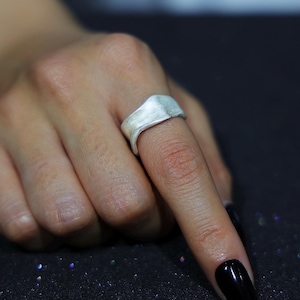 May include: A silver ring with a textured, rectangular top is worn on a finger. The ring has a smooth, curved band and a slightly irregular surface. The background is dark, with a few specks of light.