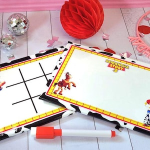 May include: Two white dry-erase boards with a black and white cow print border and yellow trim. One board has a tic-tac-toe grid, the other has the text "Samantha Story 3" and a cartoon character. A red and white marker is in front.