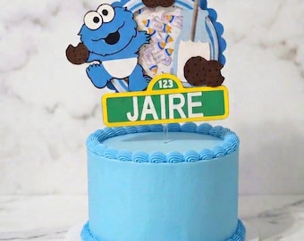 May include: A light blue frosted cake topped with a decorative cutout featuring a blue monster character, chocolate chip cookies, a milk bottle, and a green street sign with the name JAIRE and the number 123 printed in white text.