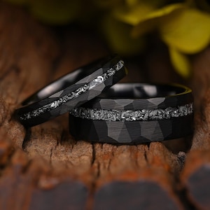 May include: Two black wedding rings with a textured, faceted surface. Each ring features a silver inlay with an intricate, patterned design. The rings are displayed on a rustic, wooden surface.