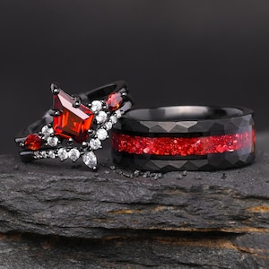 Può includere: Set di fedi nuziali nere. Un anello presenta una grande gemma rossa quadrata con pietre rosse e bianche più piccole. L'altro anello è nero con un intarsio rosso strutturato. Gli anelli sono su uno sfondo scuro.