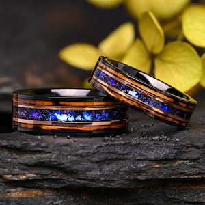 Puede incluir: Dos anillos negros con un diseño de incrustaciones de vetas de madera y cobre. La incrustación presenta un patrón moteado azul y morado. Los anillos se exhiben sobre una superficie oscura y texturizada con follaje amarillo en el fondo.