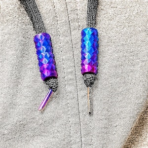 May include: Close-up of two blue and purple ombre cord ends with a geometric pattern. The cord ends are attached to black cords and have silver metal tips. The background is a grey fabric.