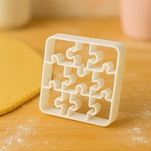 Puede incluir: Un cortador de galletas blanco con forma de rompecabezas. El cortador es cuadrado con una cuadrícula de formas de piezas de rompecabezas. El cortador está sobre una superficie de madera de color marrón claro, con masa amarilla y otros objetos en el fondo.