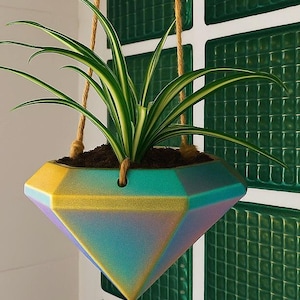 Puede incluir: Una maceta colgante geométrica con un acabado iridiscente en tonos amarillos, turquesas y morados. La maceta contiene una planta araña verde y blanca y está suspendida por una cuerda natural.