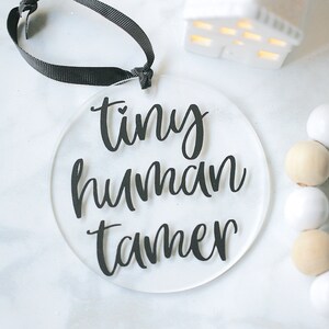 Clear 4" round acrylic ornament with black vinyl reading tiny human tamer. Hangs from black ribbon.