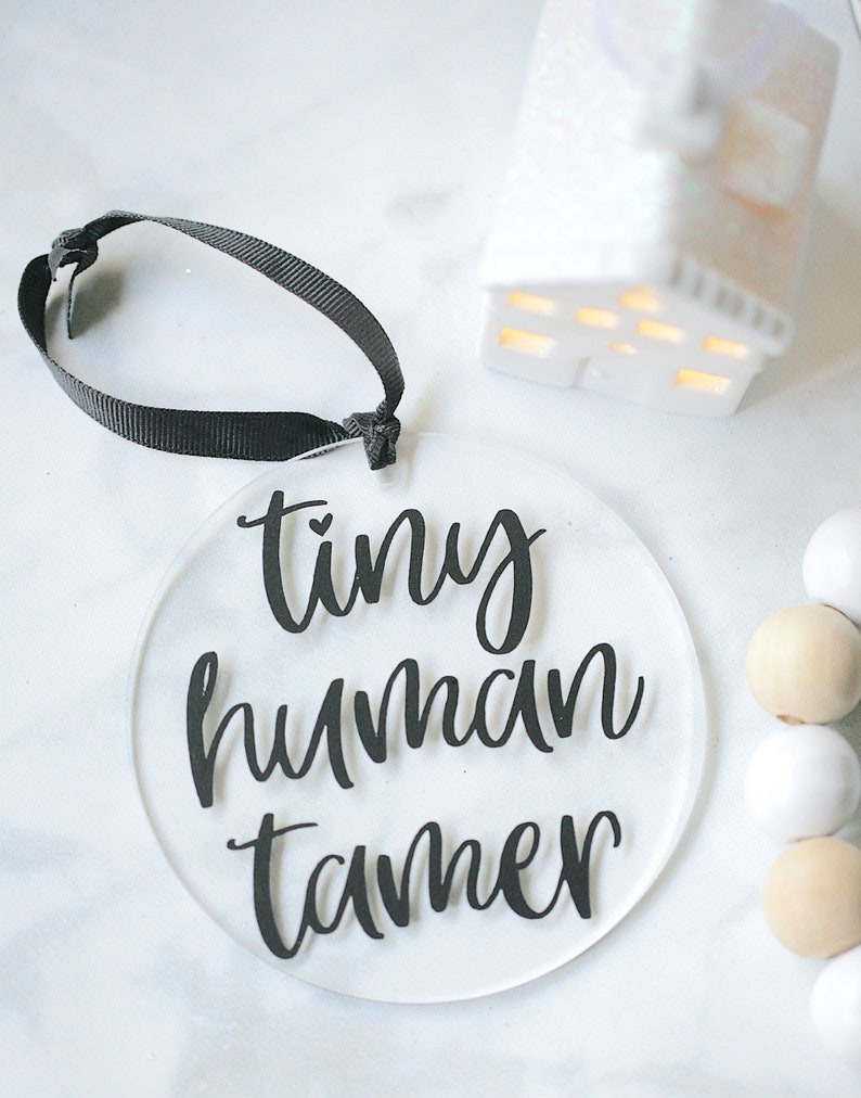 Clear 4" round acrylic ornament with black vinyl reading tiny human tamer. Hangs from black ribbon.