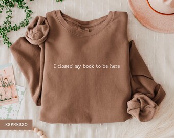 May include: Espresso-colored sweatshirt with the text "I closed my book to be here" in white. The long-sleeved top is made of a soft material and has a crew neck. The sleeves are rolled up. The word "ESPRESSO" is in a brown rectangle.