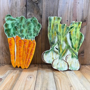 May include: Two ceramic wall plaques shaped like vegetables. The left plaque features a cluster of bright orange carrots with vibrant green leafy tops. The right plaque depicts a bunch of pale green spring onions with long, slender stalks.