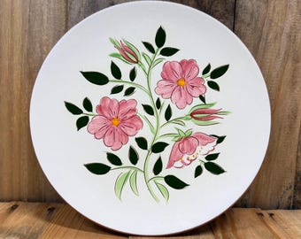 May include: A white ceramic plate with a floral design. The design features pink flowers with yellow centers, green leaves, and buds. The plate has a simple, elegant design, perfect for serving food.