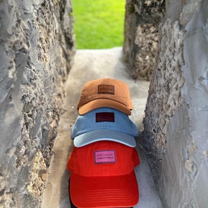 May include: A stack of four baseball caps in shades of brown, blue, and red. The top cap is brown with a rectangular patch. The second cap is light blue. The third cap is red with a pink patch that reads "LIVE, LAUGH, LOBOTOMIES".