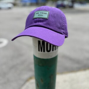 May include: A purple baseball cap with a curved brim. The cap features a rectangular patch with the text "ME TIENE MAL" in white letters on a light green background. The cap is resting on a green and white pole.