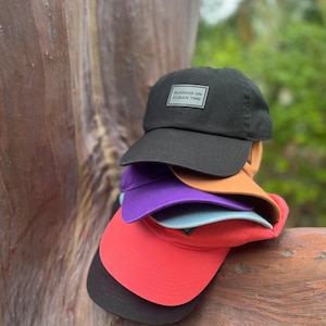 May include: A stack of baseball caps in various colors, including black, red, purple, and orange. The top black cap has a rectangular patch that reads "RUNNING ON CUBAN TIME". The caps are stacked on a wooden surface.
