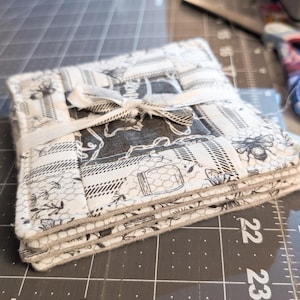 May include: A stack of square fabric coasters tied with a white ribbon. The coasters feature a patchwork design with black and white patterns, including bees, floral motifs, and honeycomb textures. The edges are a light grey.