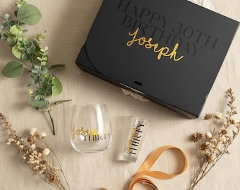 May include: A black gift box with the text HAPPY 30TH BIRTHDAY Joseph in gold and black, a stemless wine glass with the text Joseph THIRTY, a shot glass with the text Joseph THIRTY, a gold ribbon, a green eucalyptus sprig, and two dried flower stems.