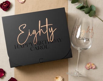 May include: A black rectangular gift box with the text Eighty HAPPY BIRTHDAY CAROL in rose gold and black lettering, accompanied by a clear glass wine goblet featuring the text Eighty CAROL in rose gold, surrounded by scattered rose petals.