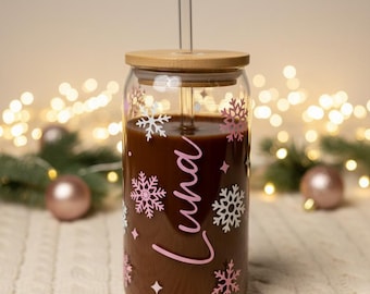 Op de afbeelding: Een doorzichtig glas gevuld met een donkerbruine drank, voorzien van een bamboe deksel en een glazen rietje. De buitenkant is versierd met witte en roze sneeuwvlokpatronen, kleine vierpuntige sterren en de naam Luna in roze schrijfletters.