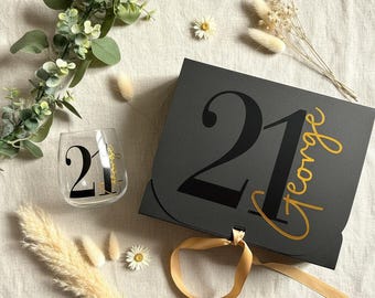 May include: A black rectangular gift box with a gold ribbon closure, featuring the number 21 and the name George in gold script. Beside it sits a clear glass tumbler with the number 21 printed in black and the name George in gold script.