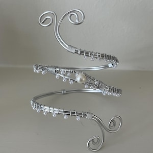 May include: A silver-toned arm cuff with a spiral design. The cuff features decorative wire wrapping and small, clear beads. A single pearl is set in the center of the design. The ends of the cuff have a curled, swirled design.