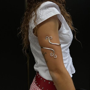 May include: A silver arm cuff with a decorative swirling design. The cuff is worn on the upper arm and features two decorative swirls. The arm cuff is a stylish accessory.
