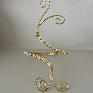 Könnte beinhalten: Goldfarbene Armspange mit Spiraldesign. Die Spange hat eine Reihe kleiner, weißer Perlen und dekorative Wirbel. Das Metall ist mit goldenem Draht umwickelt. Die Armspange wird auf einer weißen Oberfläche präsentiert.