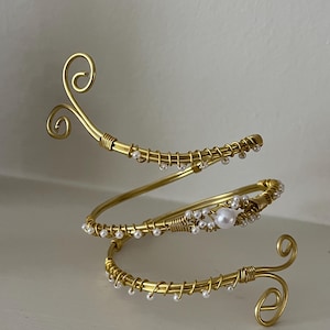Peut inclure: Un bracelet de bras doré en spirale. Le bracelet est orné de petites perles blanches et présente des volutes décoratives aux extrémités. Le bracelet est fait de fil de fer et est conçu pour être porté sur le haut du bras.