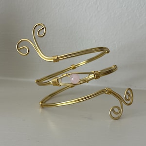 May include: Gold-coloured arm cuff with a spiral design. The cuff features a small, round, pink gemstone at the centre. The ends of the cuff have decorative swirls. The jewellery is displayed on a white surface.