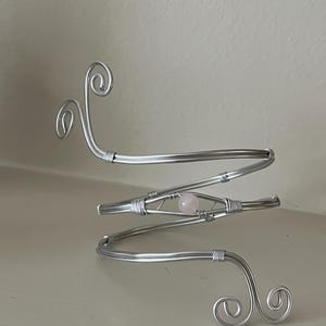 May include: A silver arm cuff with a spiral design and a small, pale pink gemstone. The cuff is made of wire and features decorative swirls at the ends. The gemstone is centered within the design.