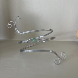 May include: Silver arm cuff with a spiral design. The cuff has a small, round, light green bead at the centre and decorative swirls at each end. The metal is wrapped around the bead and at the joints.