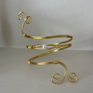 Könnte beinhalten: Ein goldfarbener Armreif mit Spiraldesign. Der Armreif hat eine kleine, runde, weiße Perle und dekorative Wirbel oben und unten. Das Metall ist zu einer Spirale gebogen, die sich um den Arm wickeln soll.