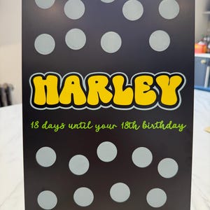 May include: A rectangular black sign featuring the name HARLEY in large yellow letters with a grey outline. Above and below the name are rows of light grey circles. The text 18 days until your 18th birthday is printed in green script.