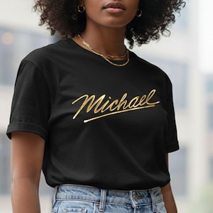 Può includere: T-shirt nero con il nome "Michael" in oro. La maglietta ha maniche corte ed è abbinata a jeans blu. È visibile anche una collana d'oro.
