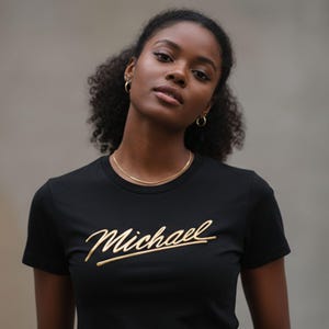 Puede incluir: Camiseta negra con el nombre "Michael" en escritura dorada. La camiseta es de manga corta y tiene cuello redondo. También se ven un collar de oro y pendientes de aro.