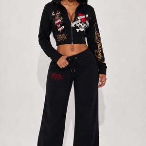May include: Black two-piece tracksuit featuring a cropped zip-up hoodie and wide-leg sweatpants. The hoodie has embroidered graphics with the text "Love Kills Slowly" and "Ed Hardy" in gold. The sweatpants also have red embroidered text.