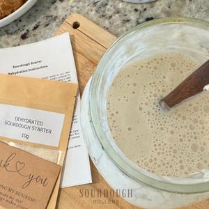May include: A glass jar with a bubbly, light-colored mixture and a wooden spoon. Beside it are instructions and a packet labeled "DEHYDRATED SOURDOUGH STARTER 10g." A small card reads "thank you" with a heart.