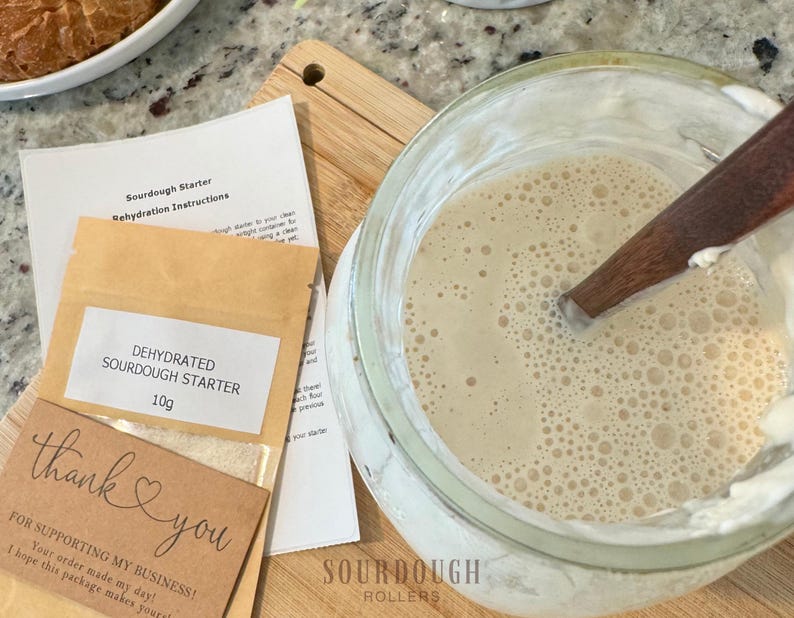 May include: A glass jar with a bubbly, light-colored mixture and a wooden spoon. Beside it are instructions and a packet labeled "DEHYDRATED SOURDOUGH STARTER 10g." A small card reads "thank you" with a heart.