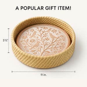Op de afbeelding: Ronde decoratieve schaal in terracotta kleur met een vogel- en bloemmotief, geplaatst in een geweven mand. De schaal heeft een diameter van 28 cm en is 9 cm hoog. De tekst "A POPULAR GIFT ITEM!" staat bovenaan.